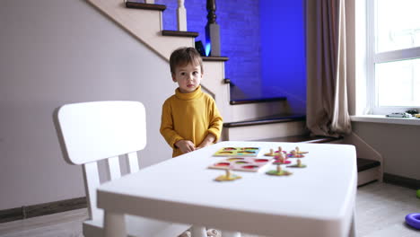 kid in yellow sweater stands near the desk. toddler turns his head to the window looking outside.