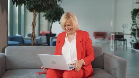 An-older-woman-in-a-red-business-suit,-sitting-on-a-couch-in-a-business-center,-watches-a-sports-match,-cheering-joyfully-at-the-victory-and-successful-bet