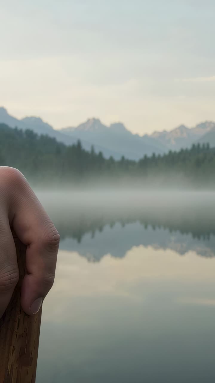 Vertical video: Pulling camera revealing mountain lake mirrored reflection, wooden paddle and mist