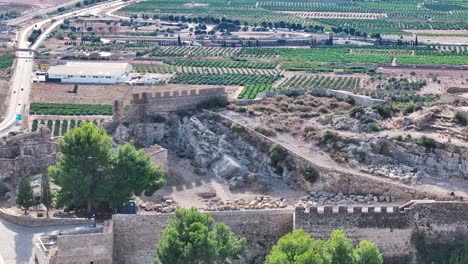 Vuelo-Con-Efecto-Paralaje-En-Un-Castillo-Vemos-Gente-Visitándolo-Y-Una-Casa-Con-Columnas-Al-Fondo-Campos-De-Cultivos-De-Naranjos-Un-Barco-Y-Una-Carretera-Con-Vehículos-Filmación-De-Alta-Calidad