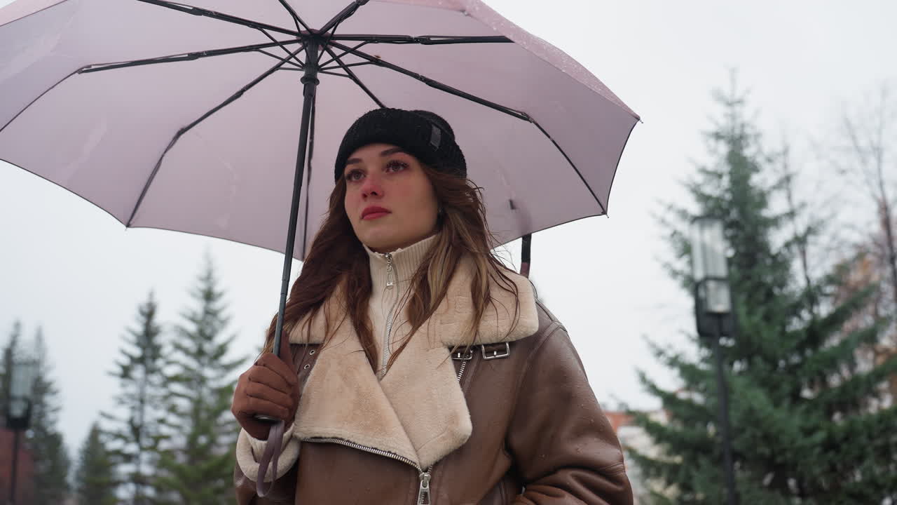 Thoughtful girl holding umbrella, wearing black knit cap and brown shearling jacket, walking calmly during cold overcast day with light snowfall, surrounded by trees and winter landscape
