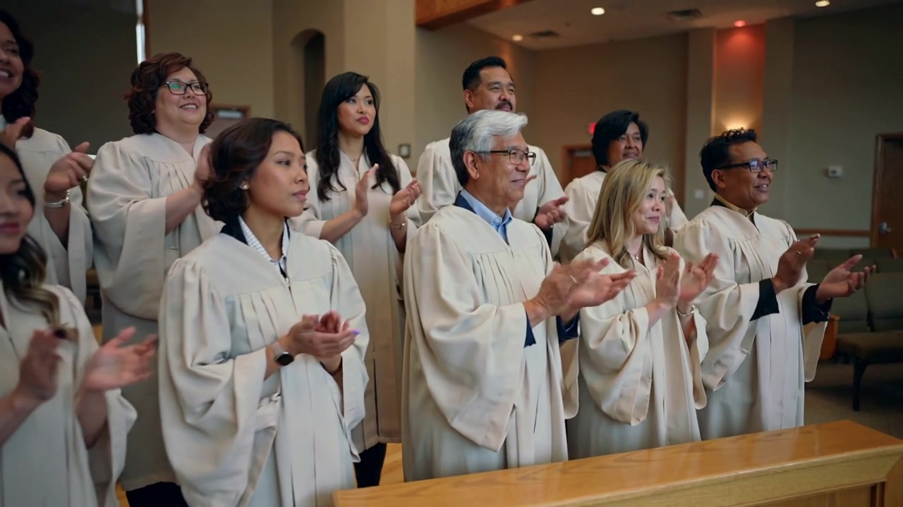 Clapping choir in beige robes responding to off-camera cue singing at chapel front by wooden rail