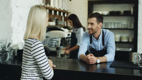 Guapo-Camarero-Hablando-Con-Una-Linda-Clienta-Rubia-En-El-Bar,-Mientras-La-Camarera-Le-Sirve-Café