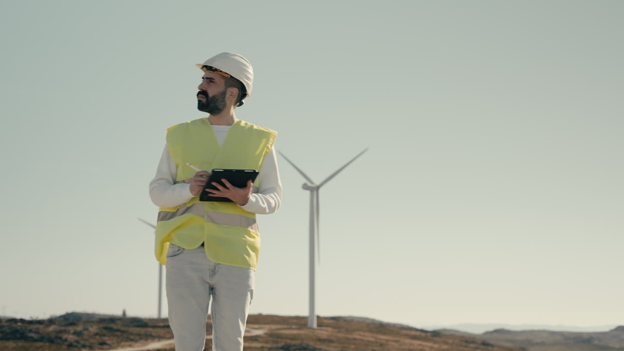 Medium shot of a focused young Caucasian male engineer using a tablet to audit wind turbines, showcasing the use of technology to support sustainable energy