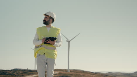 medium shot of a focused young caucasian male engineer using a tablet to audit wind turbines, showcasing the use of technology to support sustainable energy