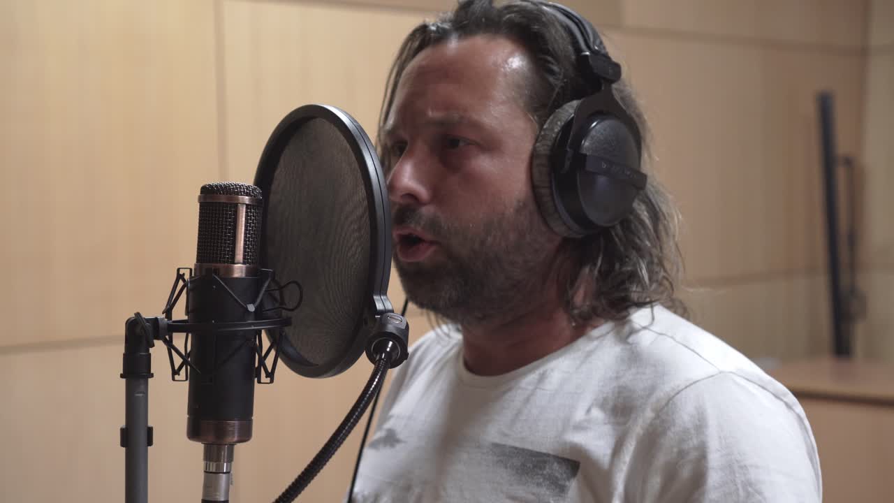 Male with long hair singing into condenser mic, recording vocals in studio, side middle shot view