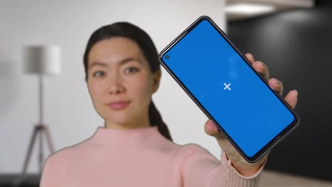 woman indoors at home in living room holding blue screen mobile phone towards camera