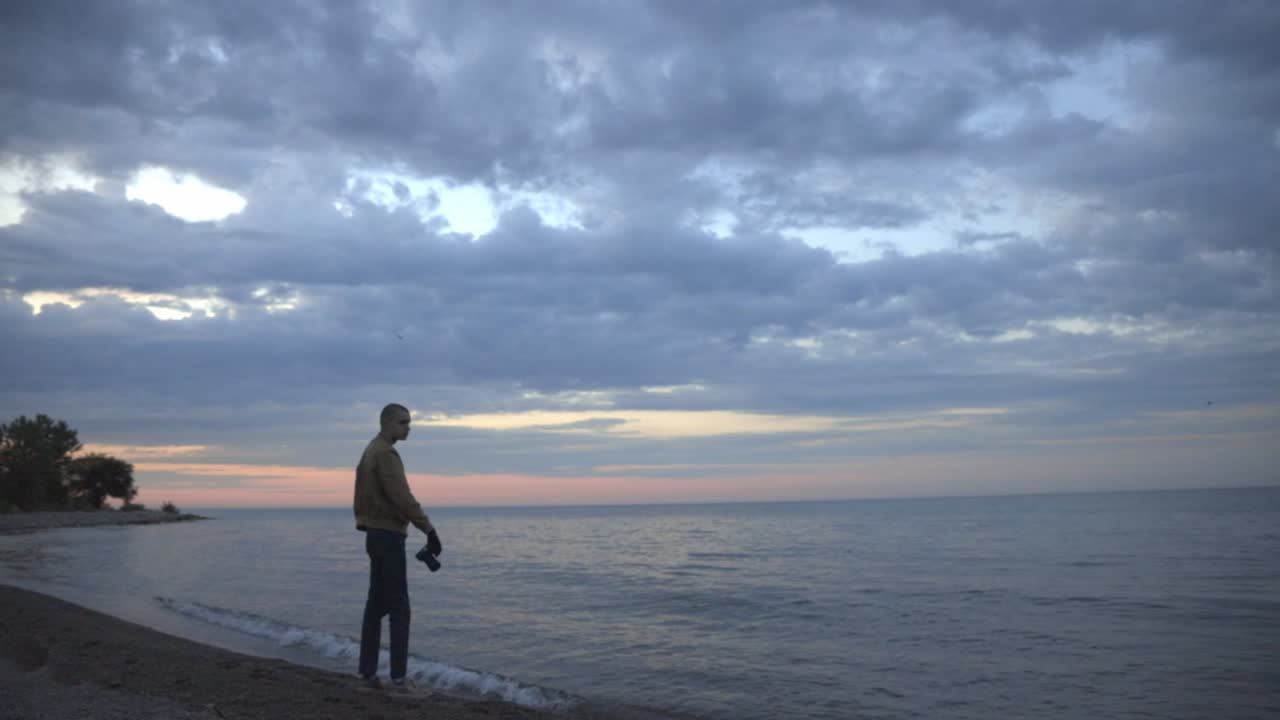 A Young Man Standing On The Shoreline While Taking Picture Of The Sunset Then Leaving During Early Evening - Wide Shot