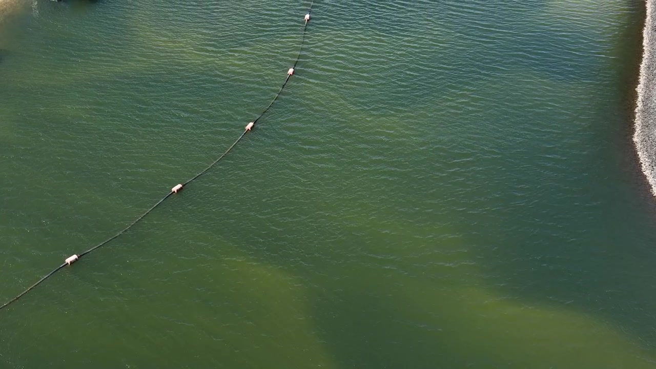 Droneoptagelse fra høj vinkel følger et langt uddybningsrør, der flyder hen over roligt grønt vand.