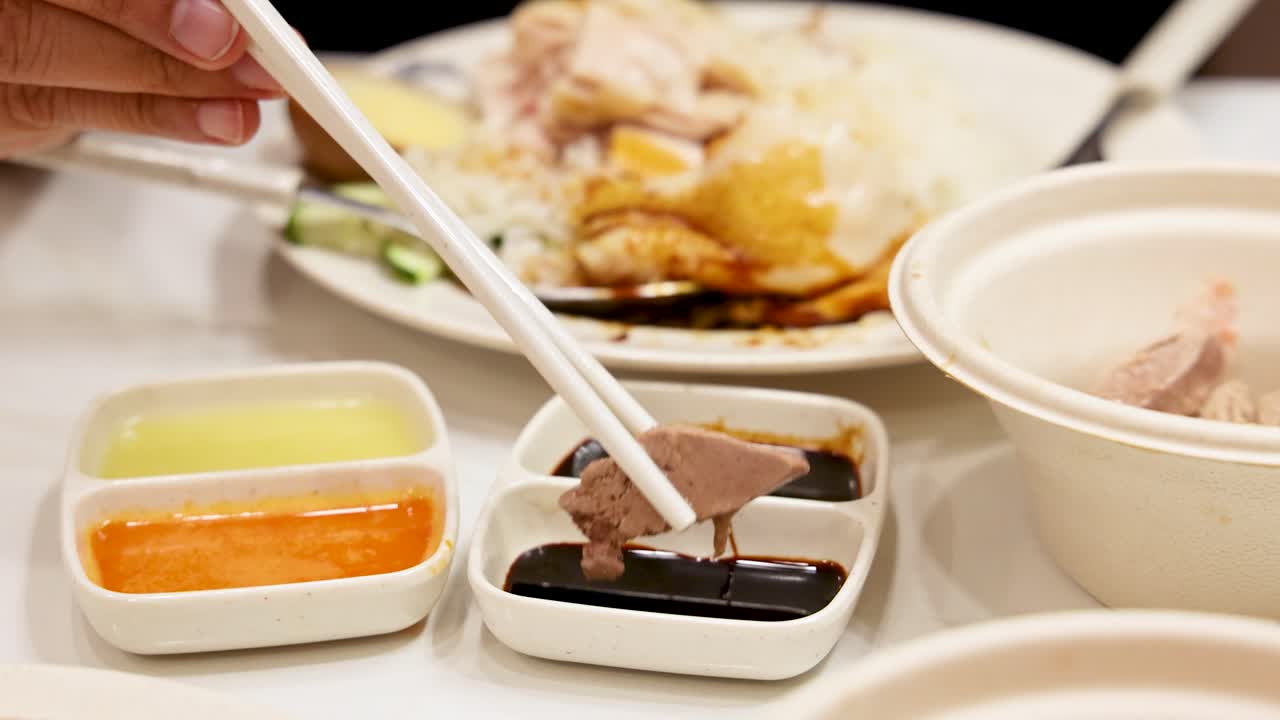 A hand uses chopsticks to dip a piece of chicken into dark soy sauce, with Hainanese rice, soup, and condiments in a bright indoor setting