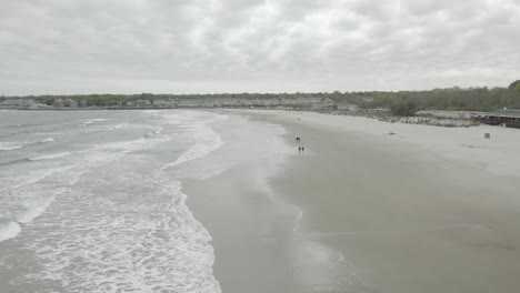 Drones-Volando-Hacia-La-Derecha-Y-Girando-Hacia-La-Izquierda,-Mientras-La-Gente-Disfruta-De-Un-Paseo-Por-La-Playa,-Clima-Nublado