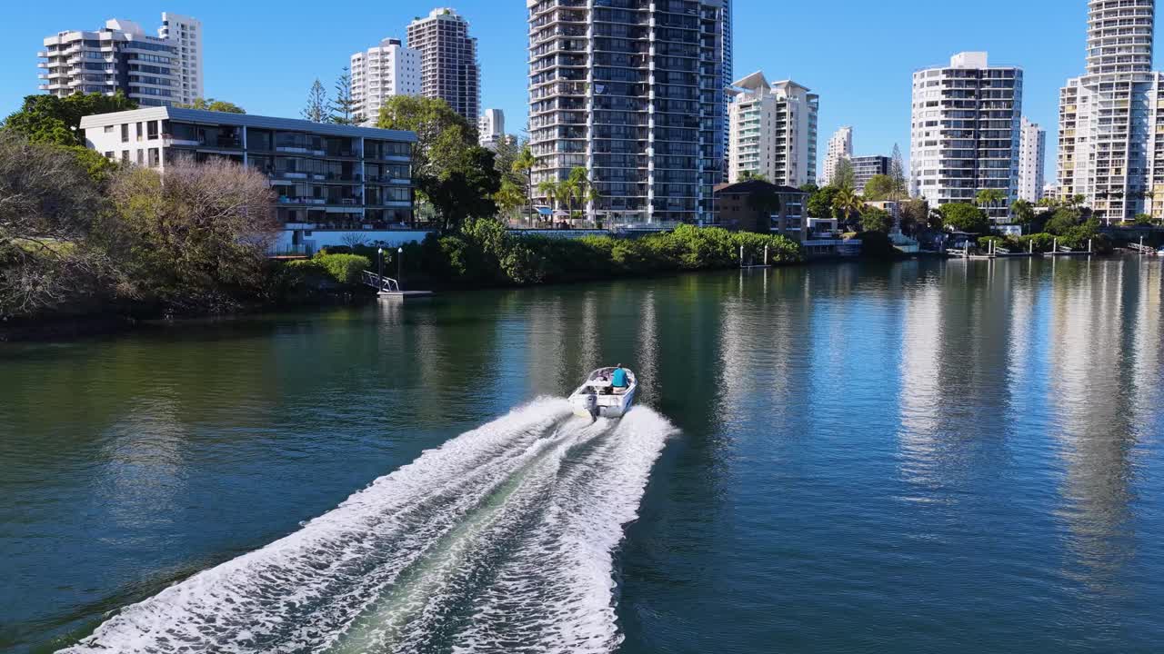 Aerial drone footage of a speedboat gliding along the Nerang River, surrounded by modern high-rise buildings under bright daylight in Gold Coast, Australia