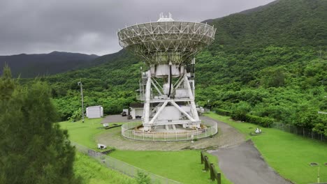 an aerial shot of a large radio telescope at a research facility, situated in a lush green valley at the base of forested mountains