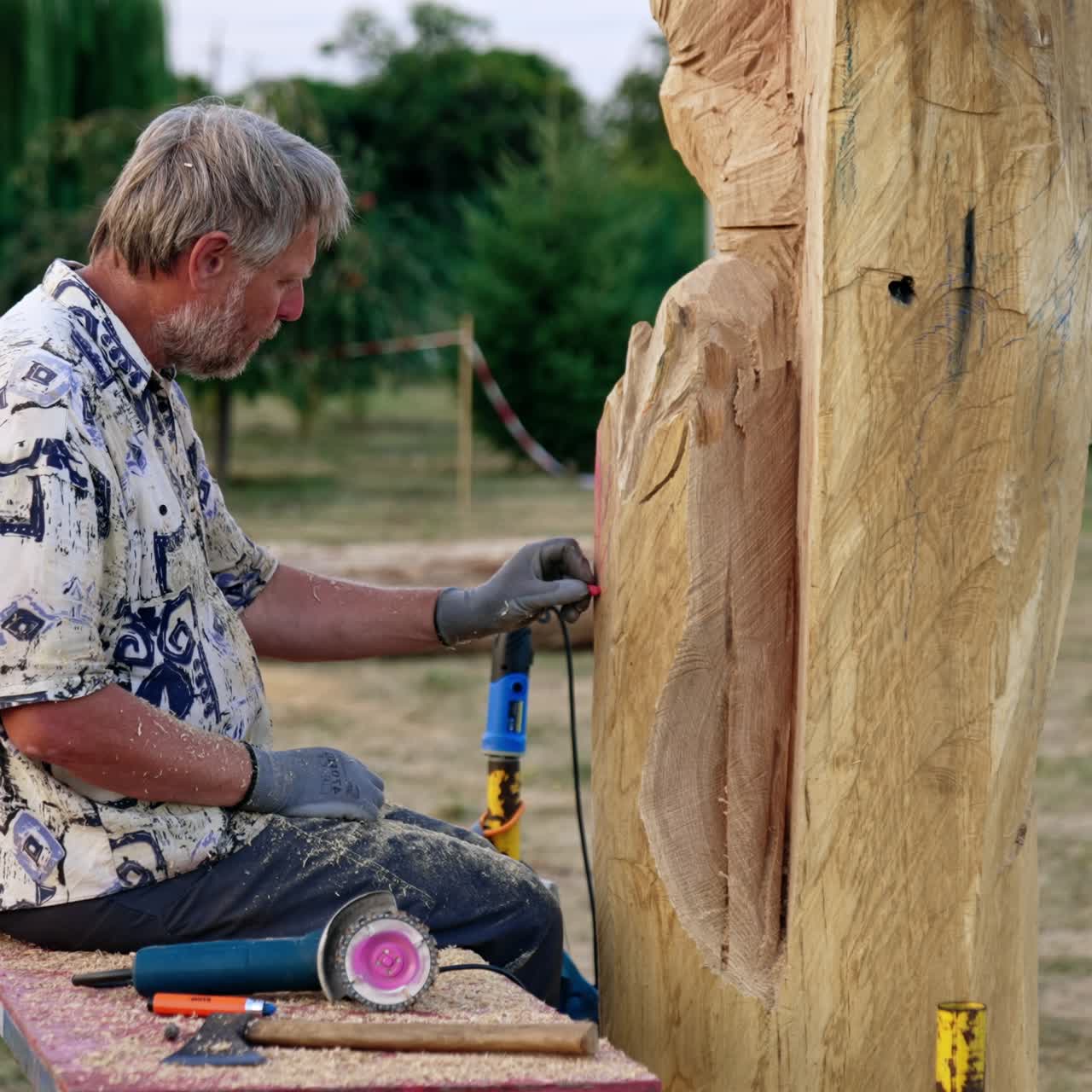 Grey-haired bearded man sits on the bench near a piece of wood outdoors. Artist draws lines on the timber with a chalk