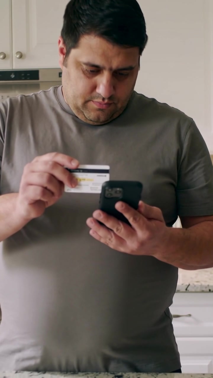 Video verticale: Uomo in maglietta grigia che guarda il telefono, allinea una carta sul bancone della cucina ed effettua un pagamento.