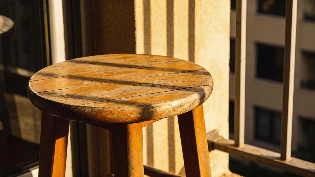 Luce solare che si sposta su uno sgabello sul balcone, spazzando le ombre delle sbarre, riscaldando la tonalità del legno per gli interni