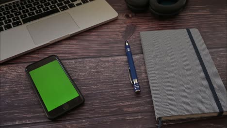 smartphone with a green screen on office wooden table with laptop, headphone - panning shot