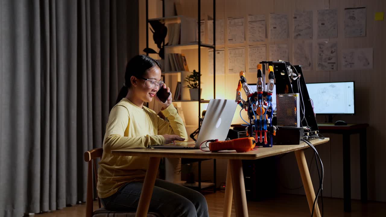 십대 아시아 소녀가 집에서 노트북에서 사이보그 손에 대해 작업하는 동안 스마트폰으로 이야기합니다.