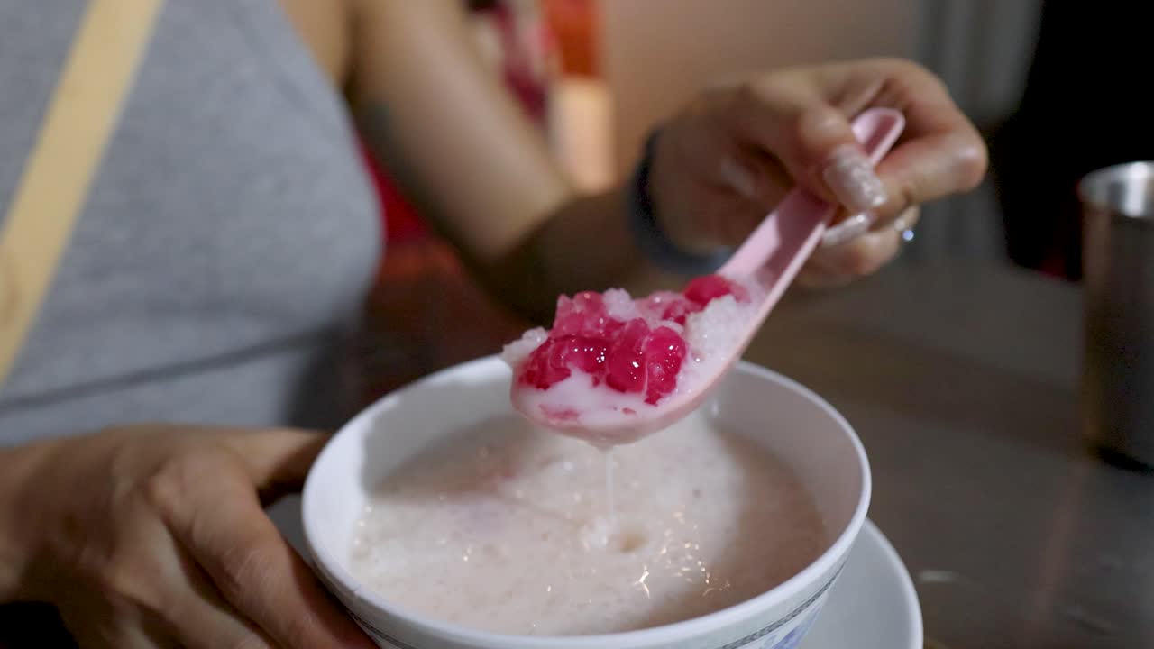 Hand lifts spoonful of red rubies dessert from bowl, street food stall, soft evening lighting