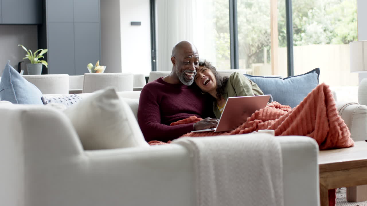 Happy biracial couple sitting on couch and using laptop at home, slow motion