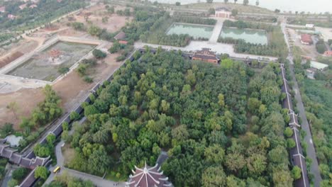 Drone-aerial-view-in-Vietnam-flying-over-a-buddhist-temple-area-filled-with-green-trees-in-front-of-a-serpent-river-in-Ninh-Binh-at-sunset-top-view