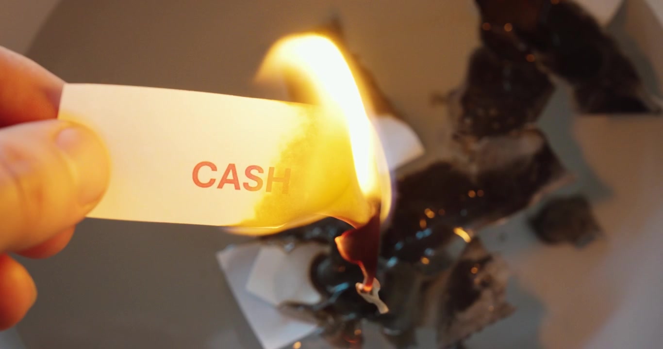 Primo piano di una striscia di carta con la parola “cash” che brucia, a simboleggiare perdite finanziarie, problemi di denaro, inflazione, rischio aziendale e pressione economica.