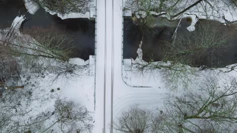 Aerial-establishing-view-of-Nordic-woodland-forest-covered-with-light-snow-and-a-dark-river-,-rural-bridge,-overcast-winter-day,-low-clouds,-birdseye-drone-shot-moving-forward