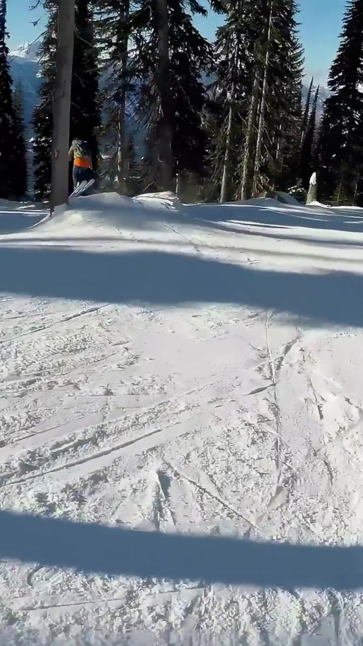Vertical video: Skiing red-jacket rider nearing bump to catch air, launching through trees on skis