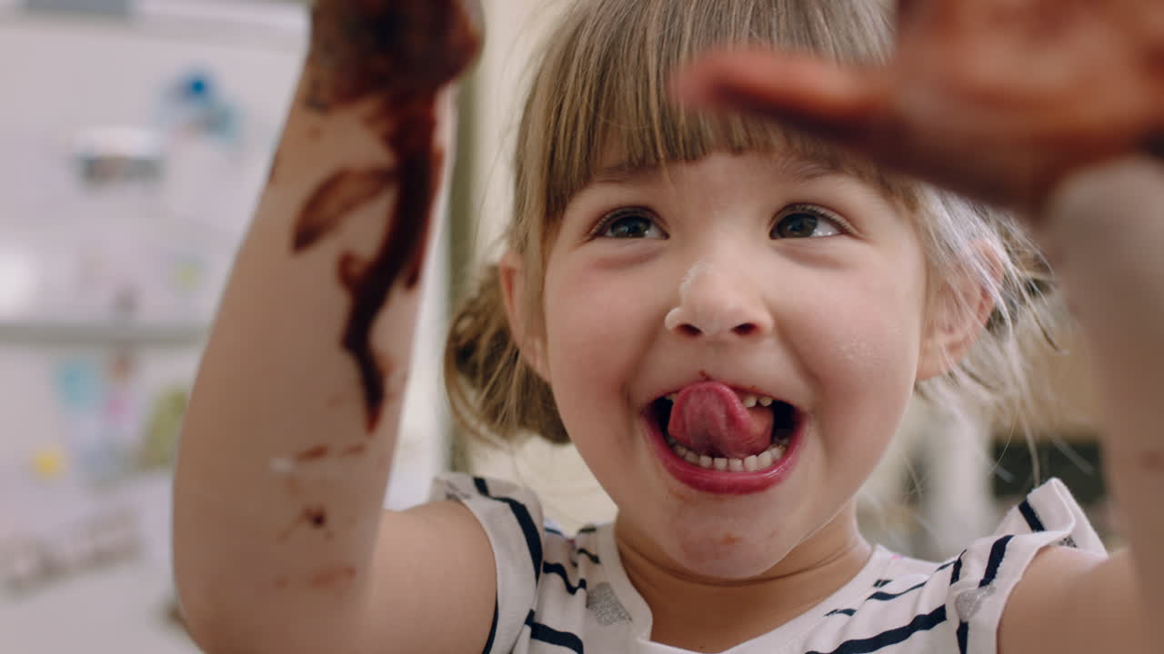 ragazza felice con le mani coperte di cioccolato leccando le dita divertendosi a cucinare in cucina bambino cattivo che gioca godendo l'infanzia