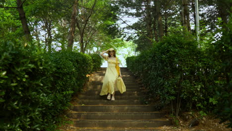 pretty woman in yellow dress holds hat, walks down tree lined stairs in park