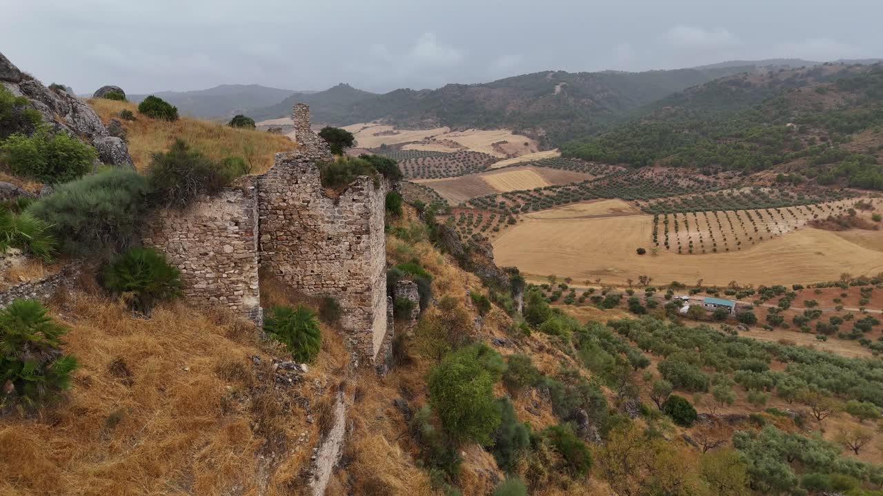 Aerial drone gliding along the side of Turón Castle ruins, Ardales, Málaga, Spain, showcasing textured stone walls and the surrounding farmland under an overcast sky