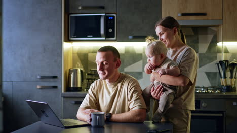 woman with baby in hands stands near her husband. man sits at the table working on laptop.