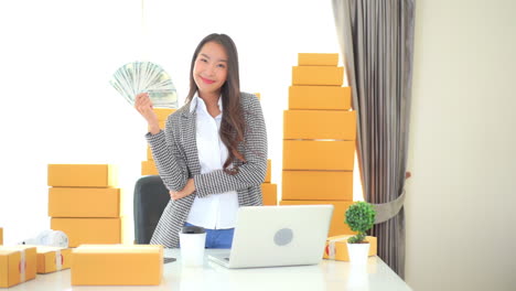 asian businesswoman working in dropshipping business holding and showing dollar banknotes money in hand standing at her workplace surrounded with yellow packages ready for shipment