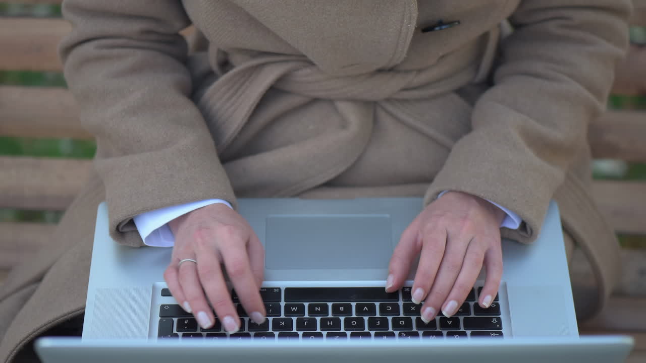 Woman in a brown coat working on her laptop on a bench in the park