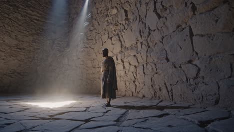 monk standing in meditative pose inside an ancient stone building, illuminated by a beam of light entering through a hole in the ceiling, creating a spiritual atmosphere