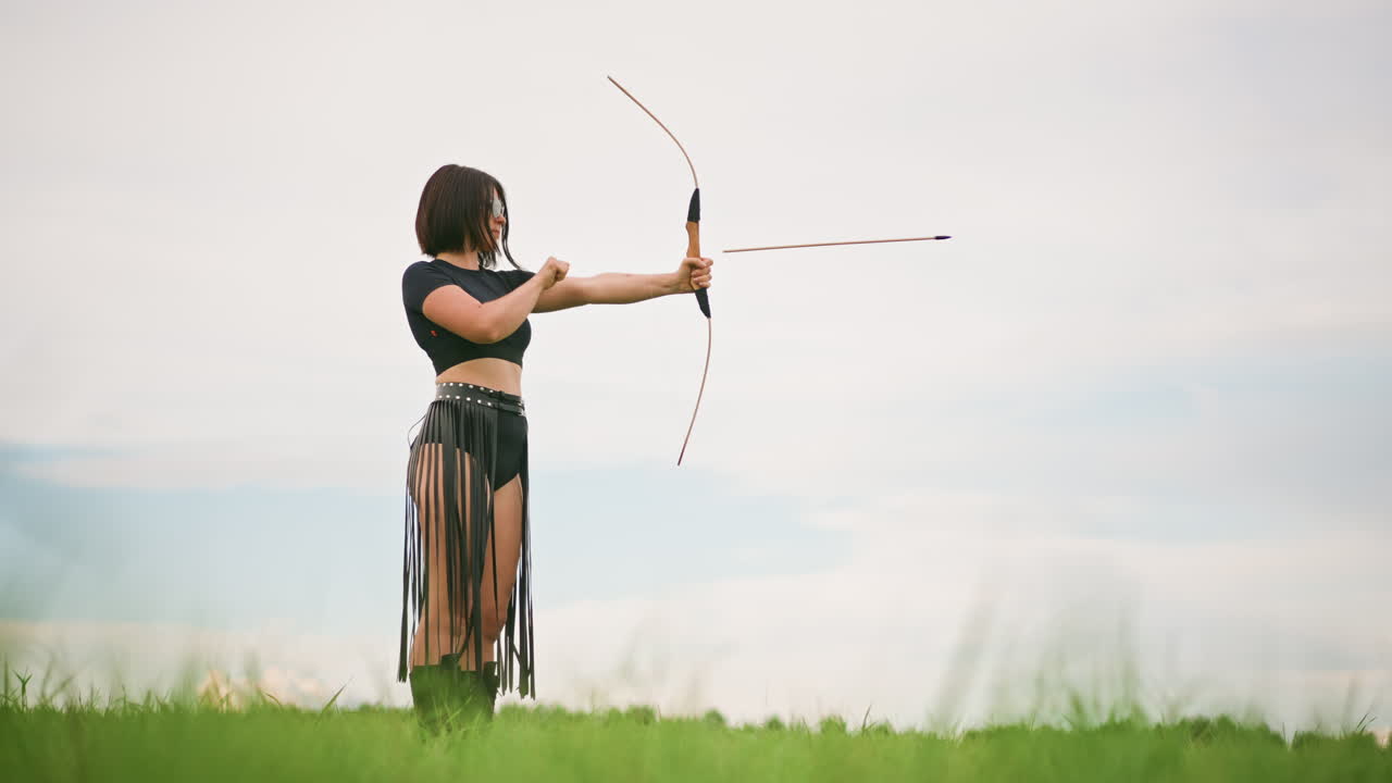 Una guerriera arciere in pelle in un campo che mira con l'arco con postura concentrata contro un cielo tenue, faretra al fianco, lunga gonna in movimento, una singola figura evoca solitudine e forza, che si esercita a tendere e rilasciare.