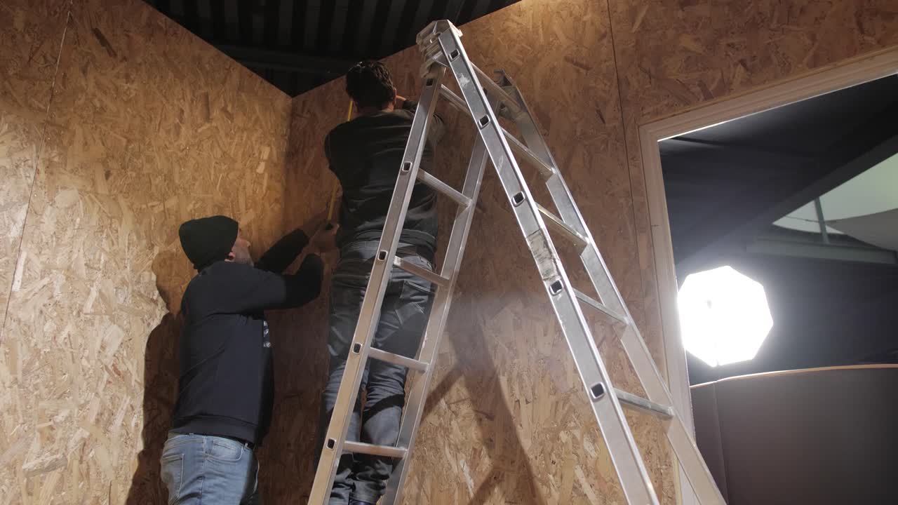 Two workers installing wall panels with ladder indoors under bright light.