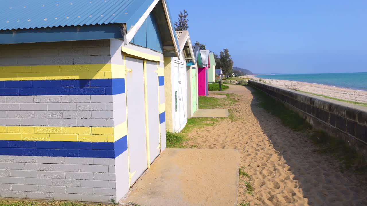 Vibrant beach huts line a sandy path under clear blue skies on Mornington Peninsula, creating a serene coastal scene