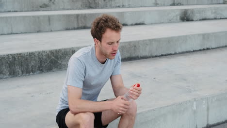 Sportlicher-Junger-Mann,-Der-Nach-Dem-Lauftraining-In-Der-Stadt-Auf-Treppen-Sitzt-Und-Wasser-Trinkt