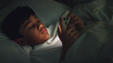 tired young boy in bedroom at home lying in bed using mobile phone to text message at night