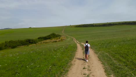 aerial follow shot of male walking on dirt path through green countryside on sunny day