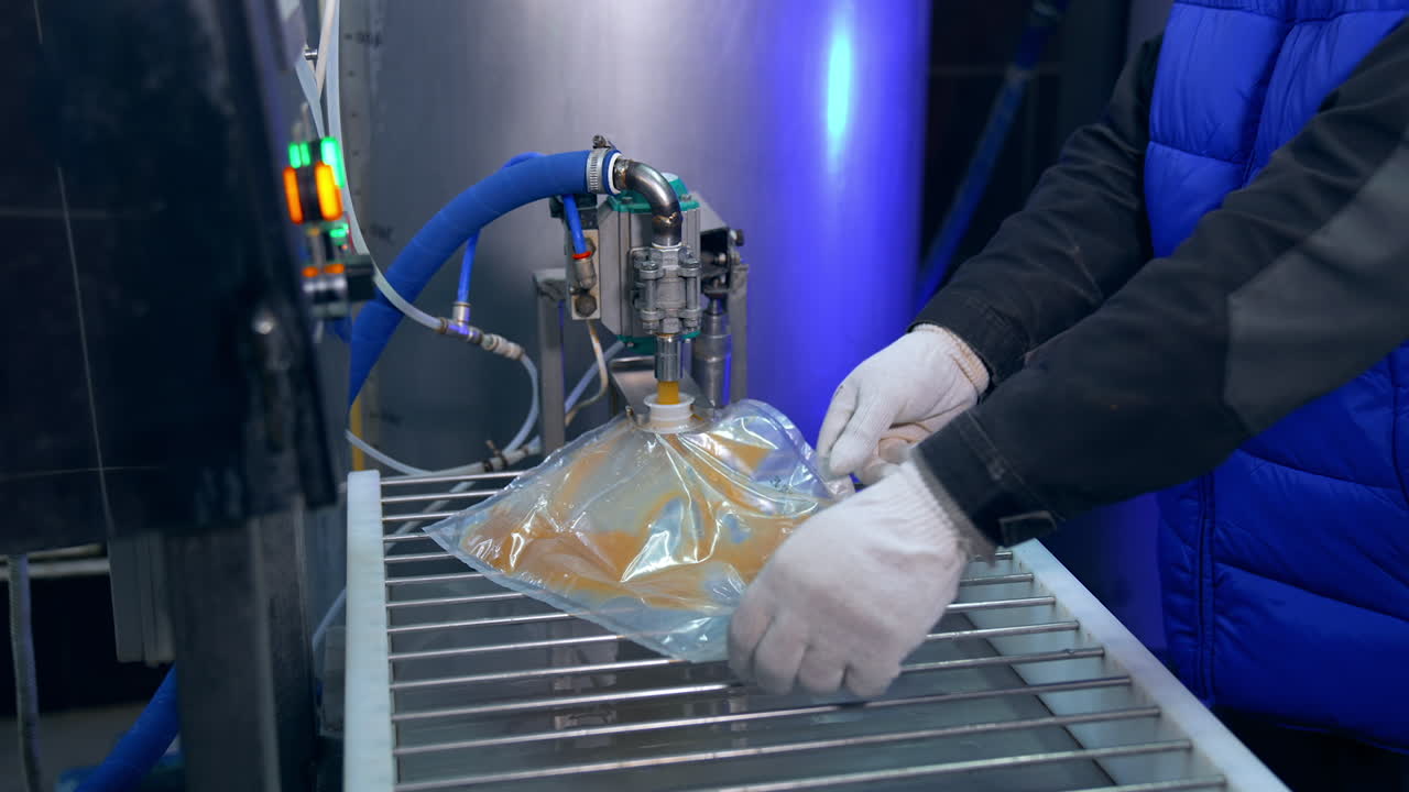 Transparent plastic bag being filled with juice through an opening. Factory worker smoothes out the bag for better filling of juice.