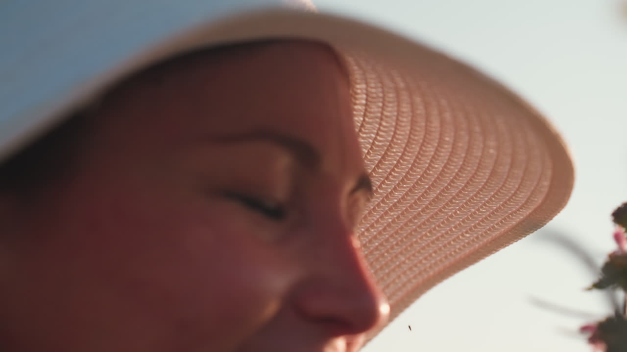 close up of lady in sun hat gently smiling with eyes closed while scenting vibrant wildflowers under soft daylight, capturing peaceful connection with nature and delicate moment of contentment