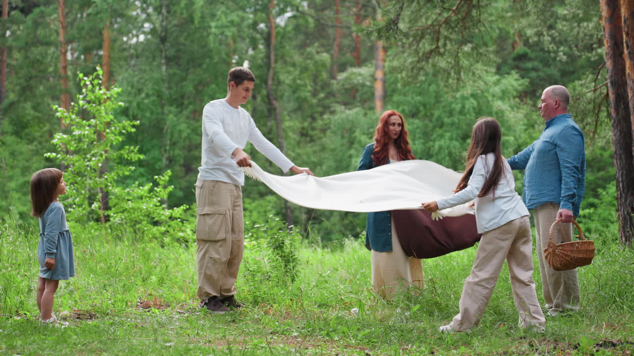 아이들이 풀밭에 우유색 천을 펼치고 부모는 갈색 천을 펼치는 동안, 아버지가 바구니를 들고 어린 소녀가 호기심을 가지고 바라보는 행복한 가족이 숲속에서 피크닉 장소를 정리하는 모습의 롱 샷