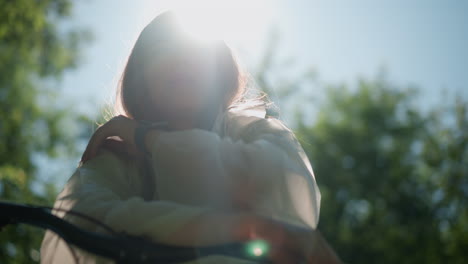 close-up of lady in translucent raincoat lowering hood with sunlight highlighting her contemplative expression, she rests her hand thoughtfully, with lush greenery softly blurred in background