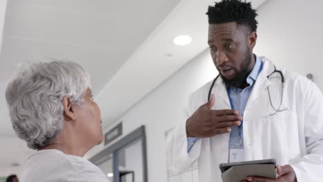 diverse serious male doctor with tablet talking to senior patient on bed in slow motion, unaltered