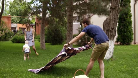 caucasian family outdoors. father, mother and little blondy son. dad spreads a picnic cover, picnic basket. green lawn