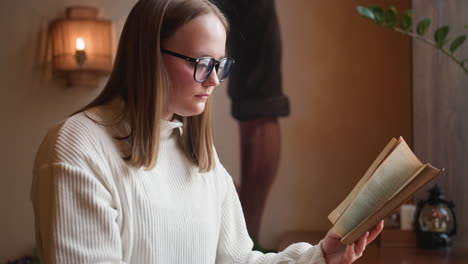 young girl wearing glasses reads book in cozy cafe setting while holding coffee mug, soft decorative light glows in background creating warm ambiance near wooden table and indoor plant by window