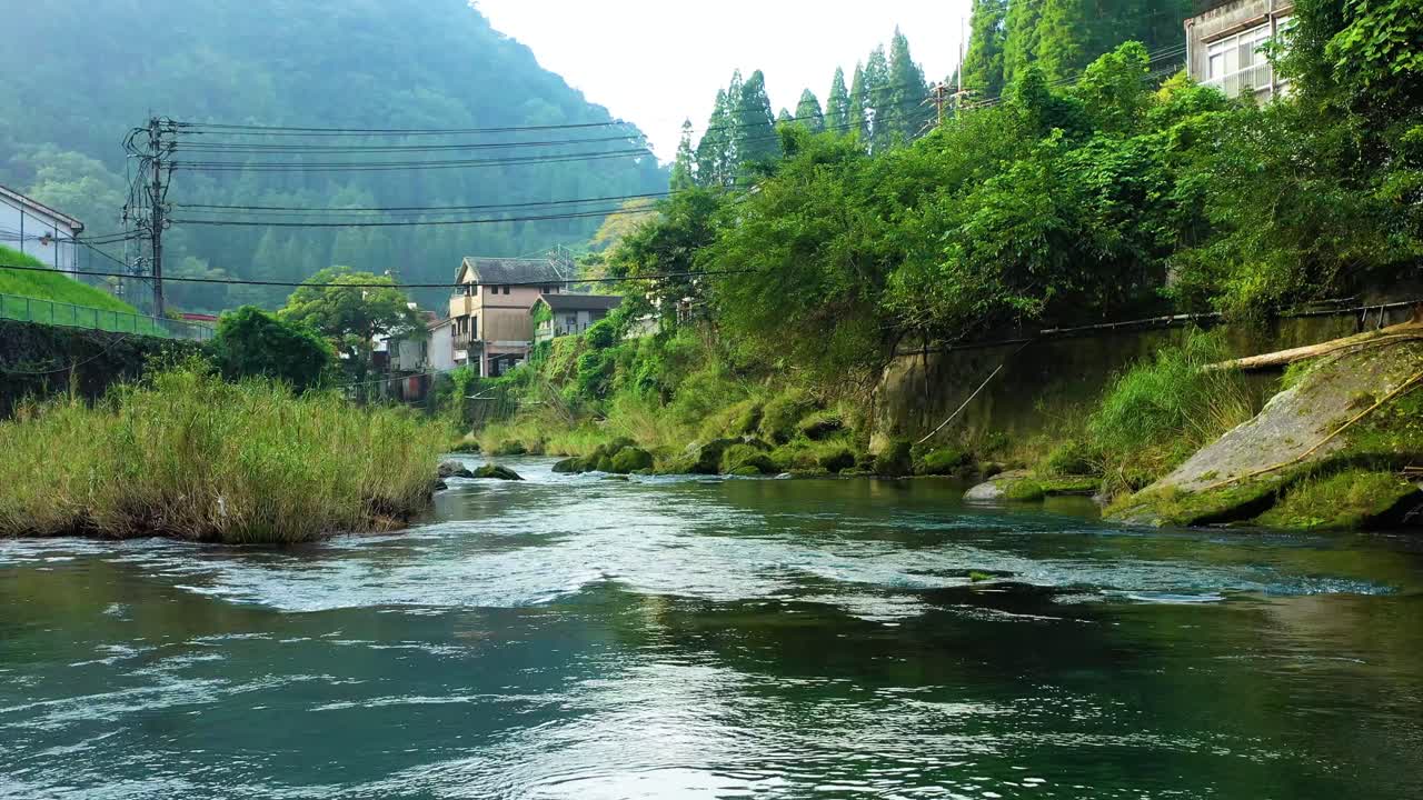 vuelo de drones sobre un río prístino en la pacífica ladera japonesa, exuberante vegetación que rodea el pintoresco pueblo