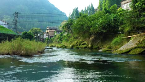 Vuelo-De-Drones-Sobre-Un-Río-Prístino-En-La-Pacífica-Ladera-Japonesa,-Exuberante-Vegetación-Que-Rodea-El-Pintoresco-Pueblo